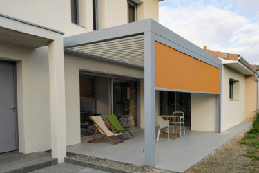 Pergola bioclimatique grise avec lames orientables sur une terrasse carrelée de maison, store orange rabattu, salon de jardin et chaises longues.
