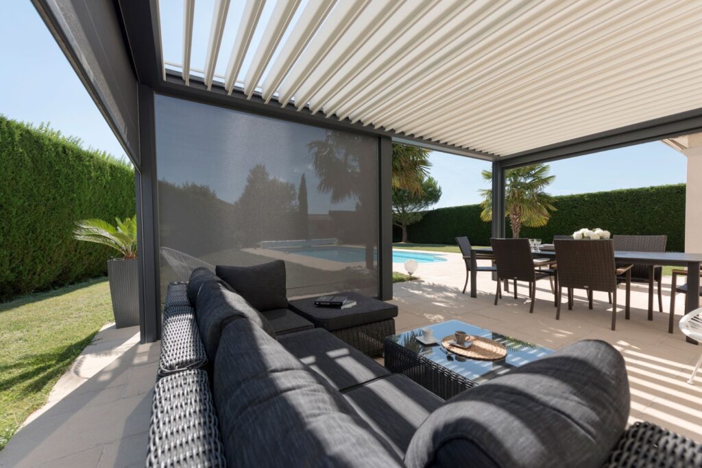 Pergola bioclimatique à lames orientables avec store latéral ombrageant un salon de jardin et une salle à manger près d'une piscine ensoleillée.