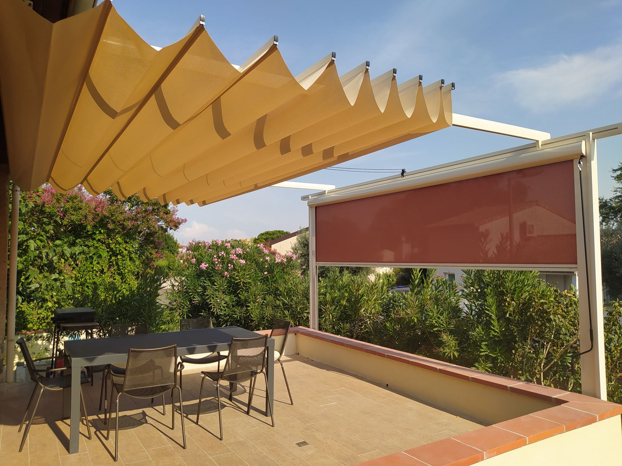 Terrasse extérieure ombragée par une pergola rétractable beige, avec table, chaises, barbecue et végétation fleurie.