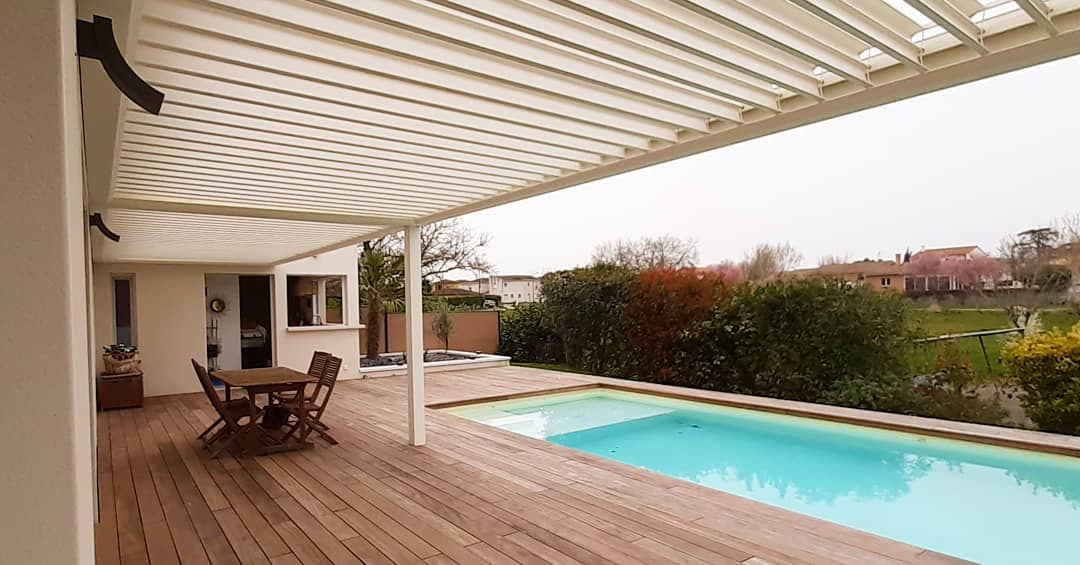 Terrasse bois avec piscine et pergola bioclimatique à lames orientables, attenante à une maison. Mobilier extérieur.