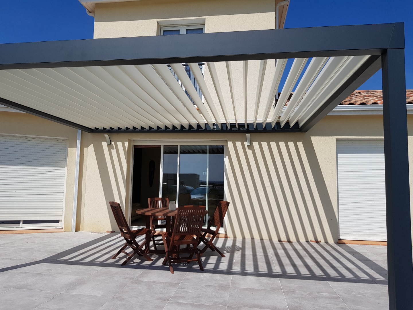 Pergola bioclimatique moderne avec lames orientables sur une terrasse carrelée, salon de jardin en bois, ombres portées.