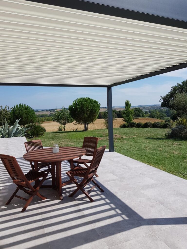 Patio avec table et chaises en bois sous une pergola bioclimatique. Ombre portée sur la terrasse carrelée. Vue sur paysage vallonné.