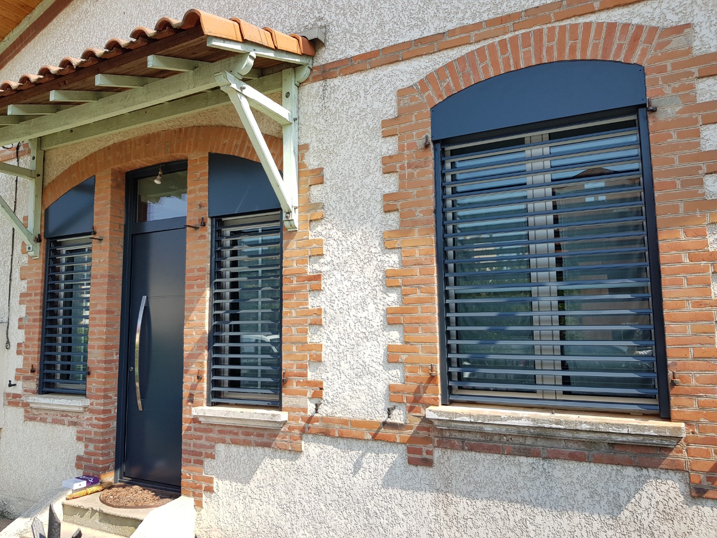 Façade maison avec porte sombre, 2 fenêtres grillagées, arches en briques, auvent bois et tuiles