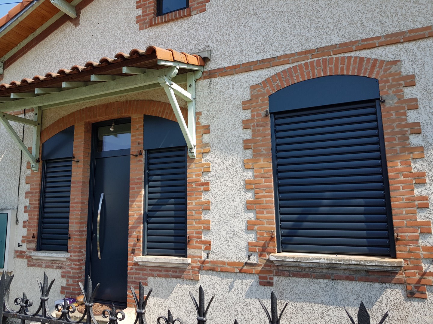 Façade maison avec porte noire, volets roulants noirs sur fenêtres à arches en brique. Auvent tuilé, mur texturé, clôture fer.
