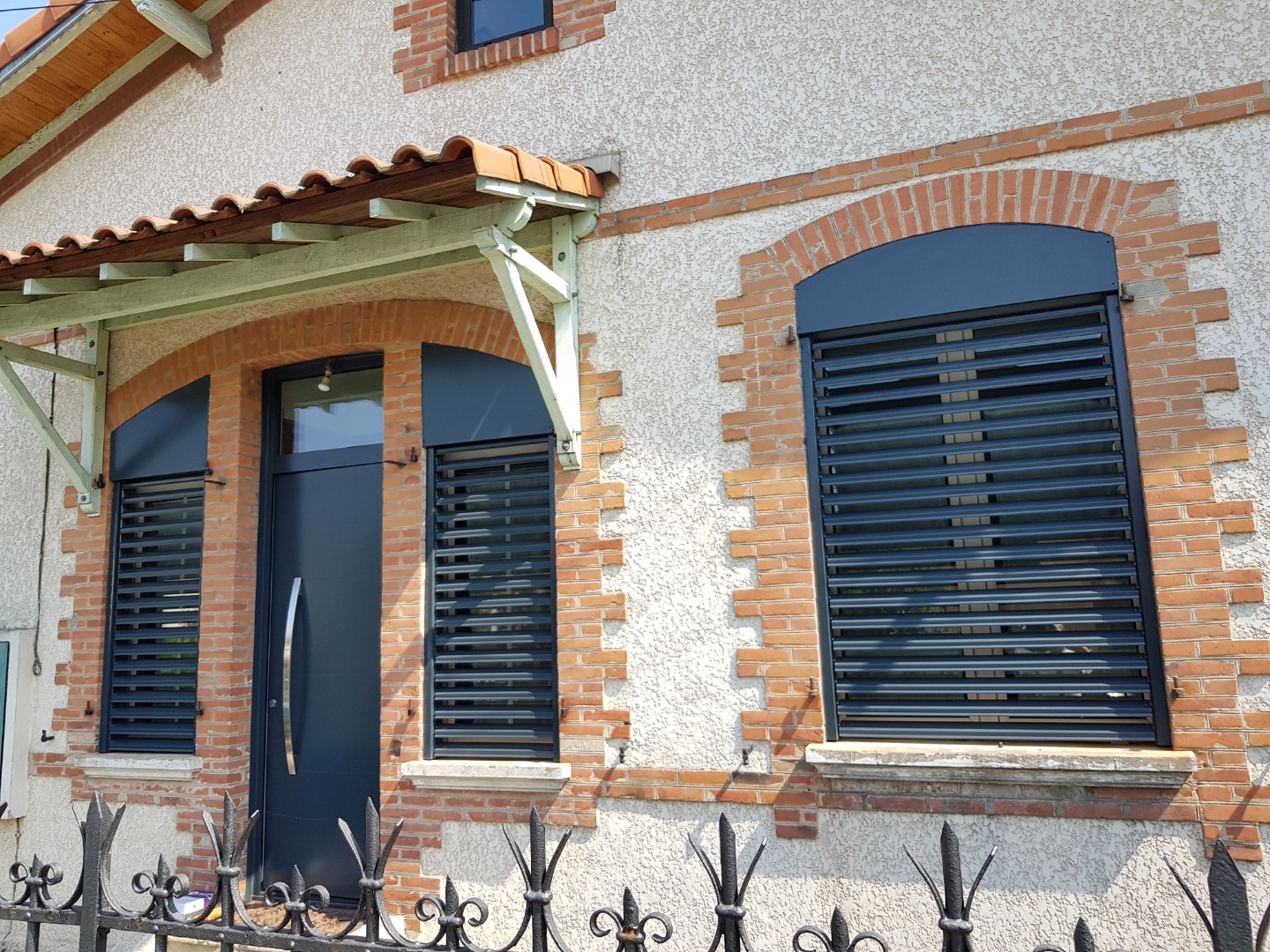 Façade de maison en briques avec volets persiennes gris modernes et porte d'entrée assortie. Auvant tuilé et clôture noire.