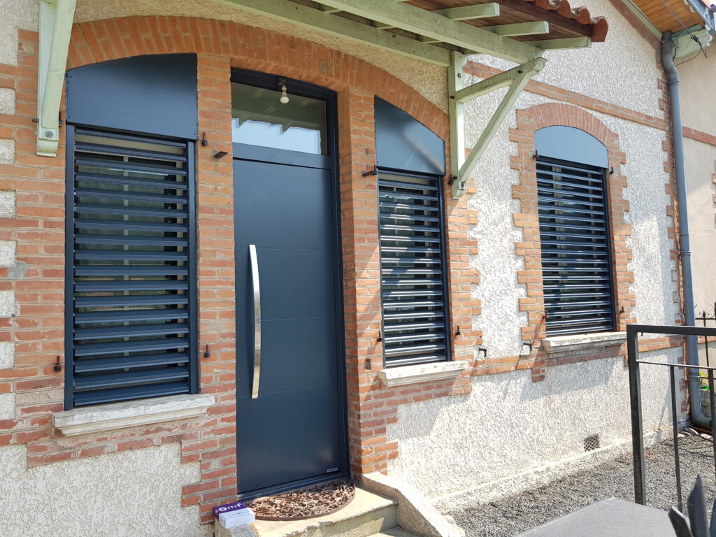 Façade maison: porte grise moderne, volets persiennes, arcades brique. Boîte Omfy sur marche d'escalier.