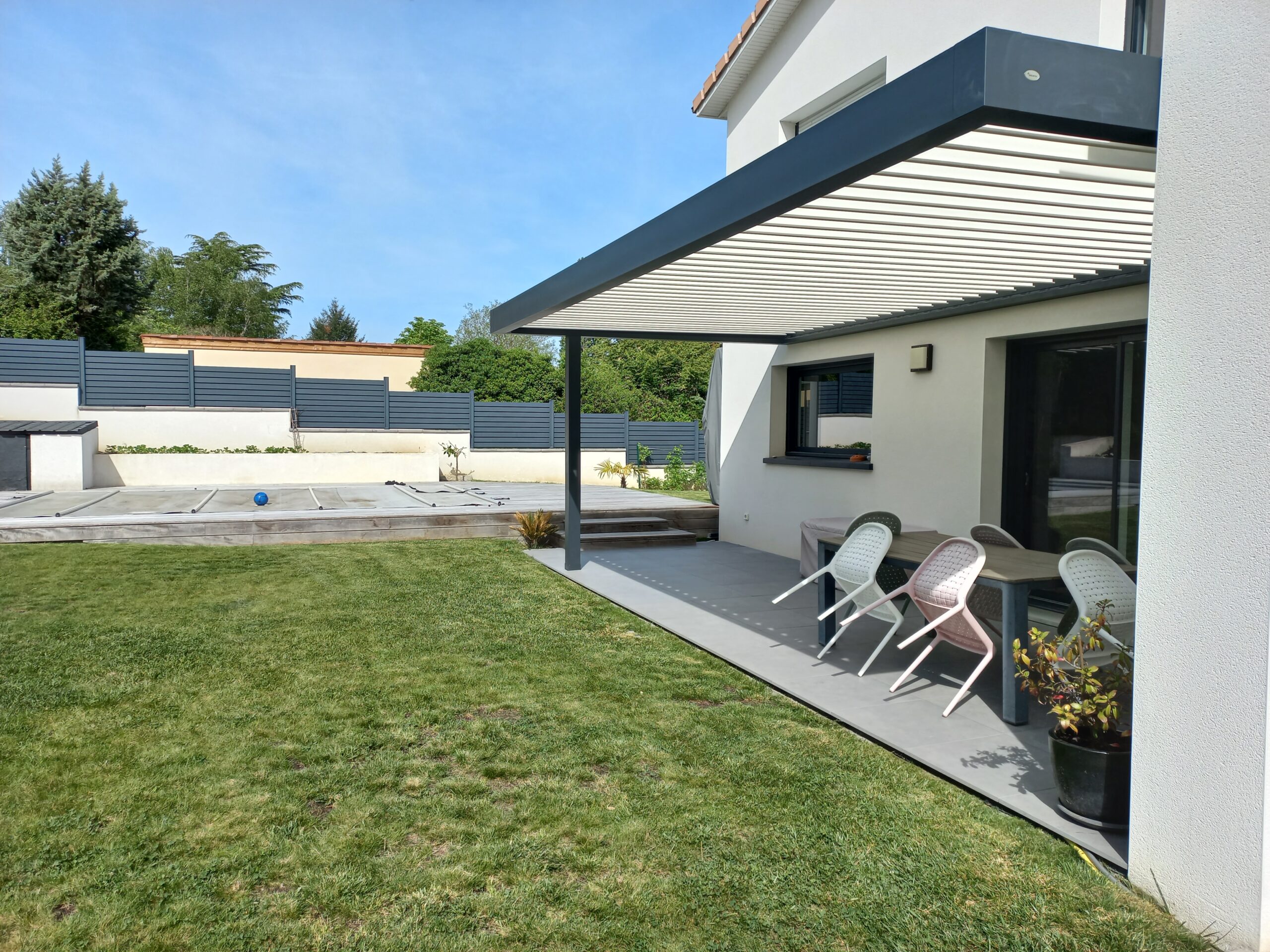 Terrasse carrelée moderne avec pergola bioclimatique lames orientables. Pelouse verte, piscine couverte, clôture grise.