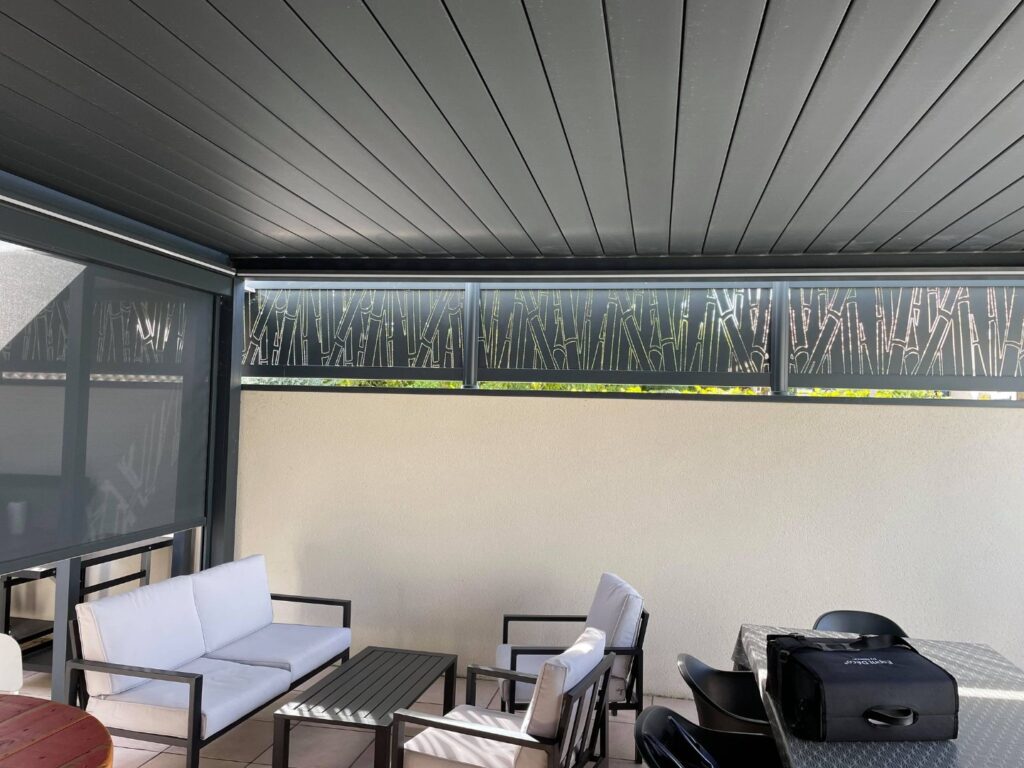 Terrasse moderne avec toit lamellé, écran rétractable, et panneaux décoratifs ajourés motif bambou, mobilier extérieur blanc et gris.