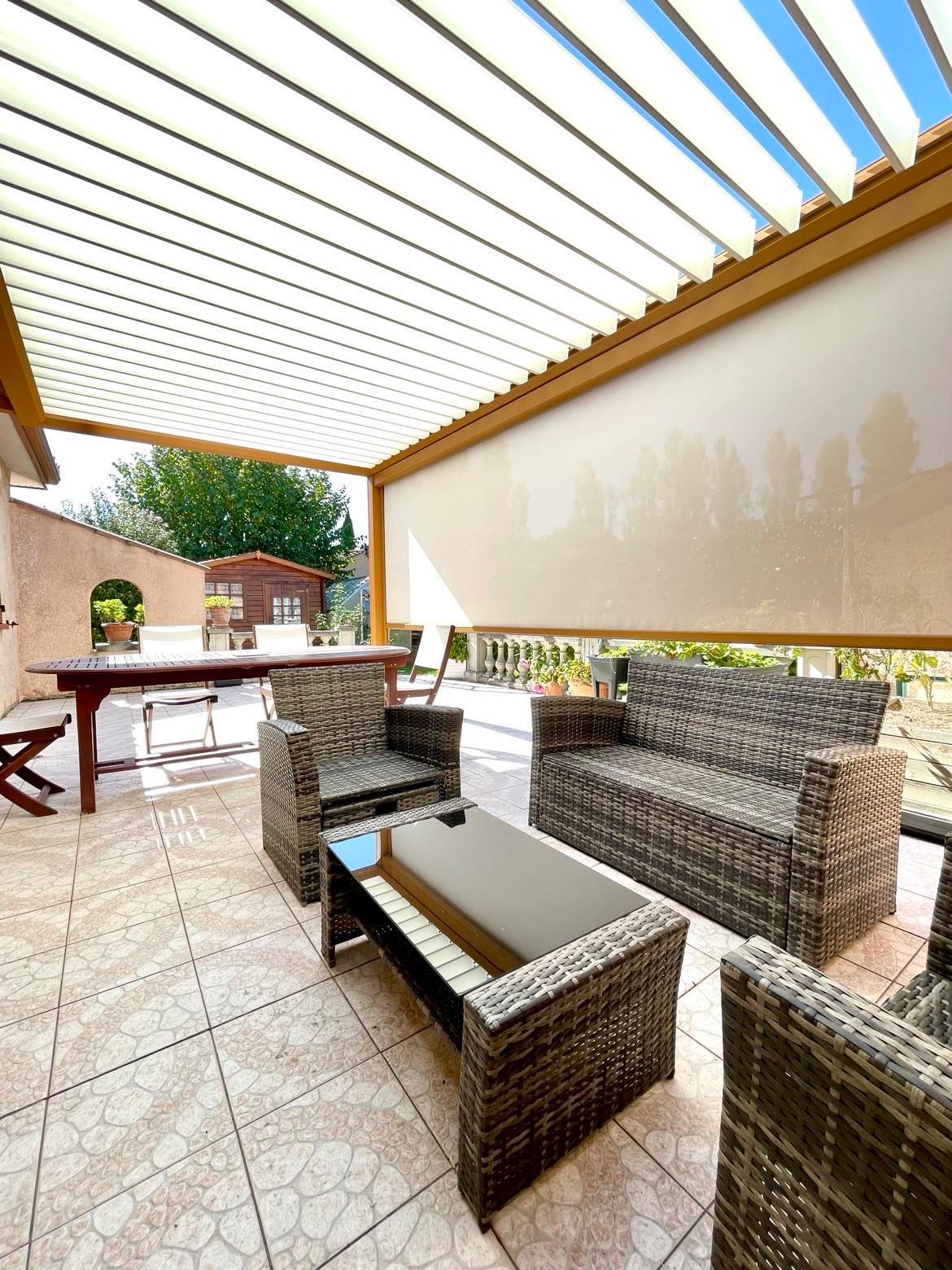 Patio carrelé sous pergola bioclimatique, avec salon de jardin en rotin et table à manger. Store latéral visible.