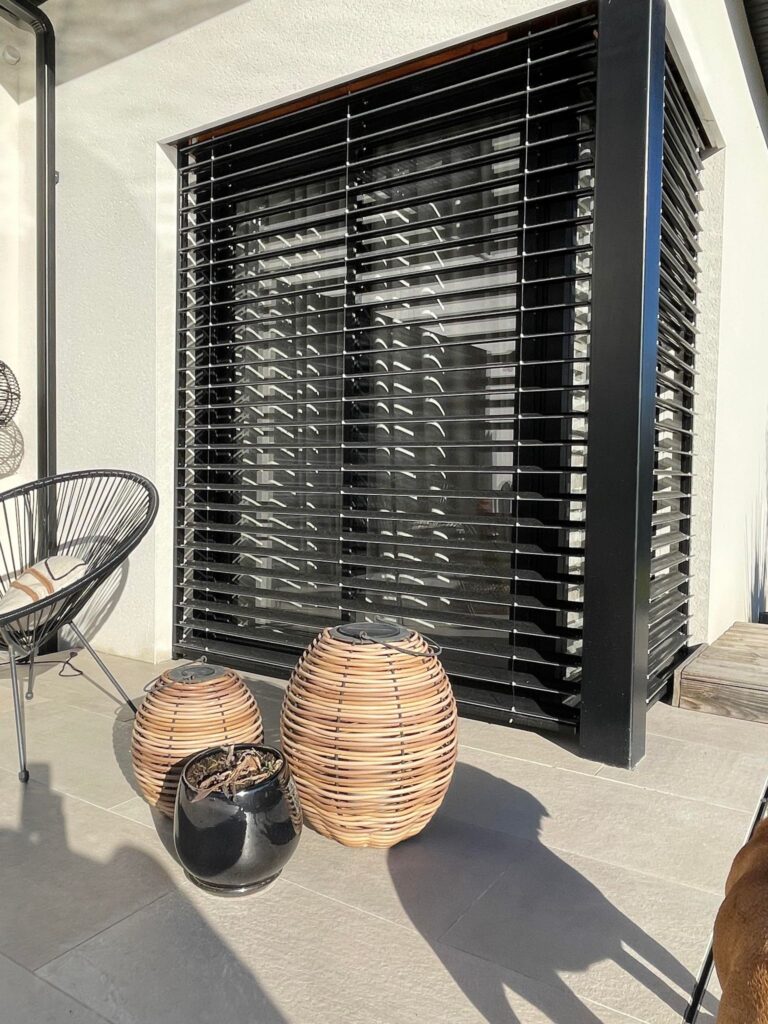 Store extérieur noir à lames sur grande fenêtre, avec deux lanternes tressées, pot noir et chaise sur terrasse ensoleillée.