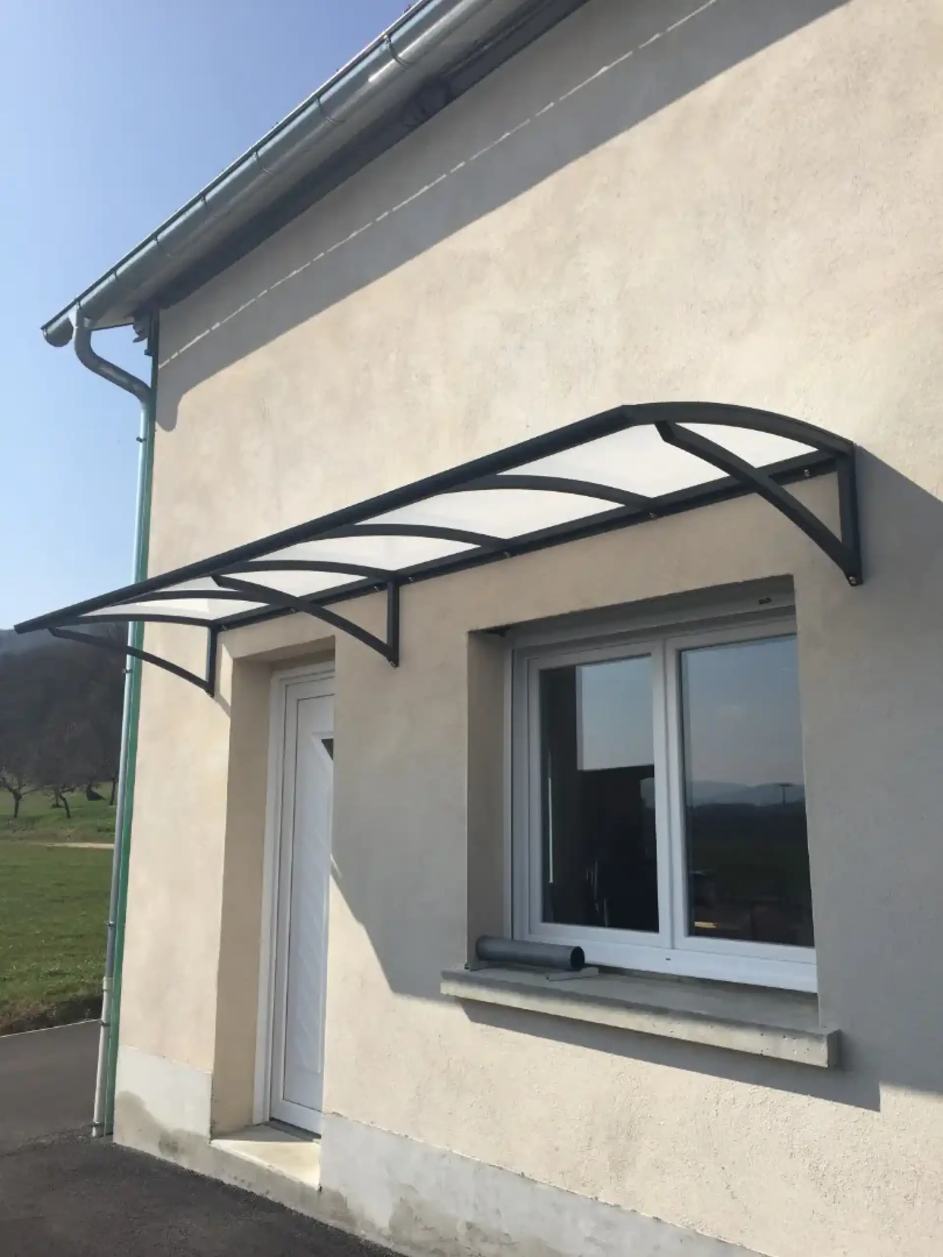 Auvent polycarbonate sur structure noire protégeant porte et fenêtre d'une façade claire de maison. Gouttière verte.