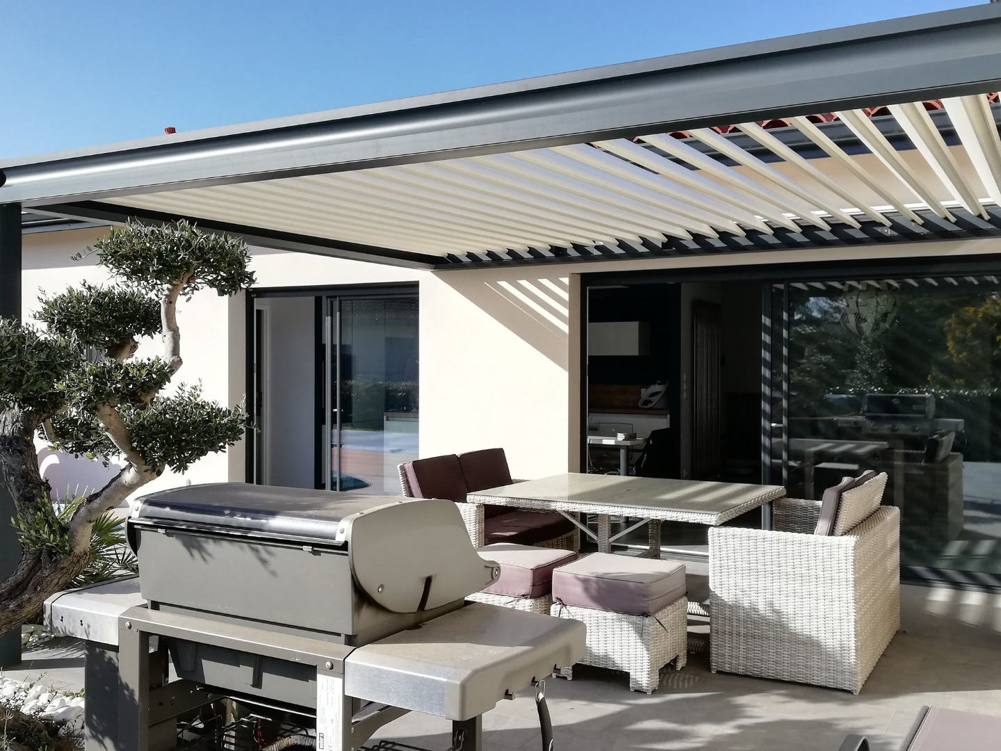 Patio moderne avec pergola bioclimatique, mobilier de jardin blanc, barbecue et olivier. Vue sur l'intérieur de la maison par une baie vitrée.