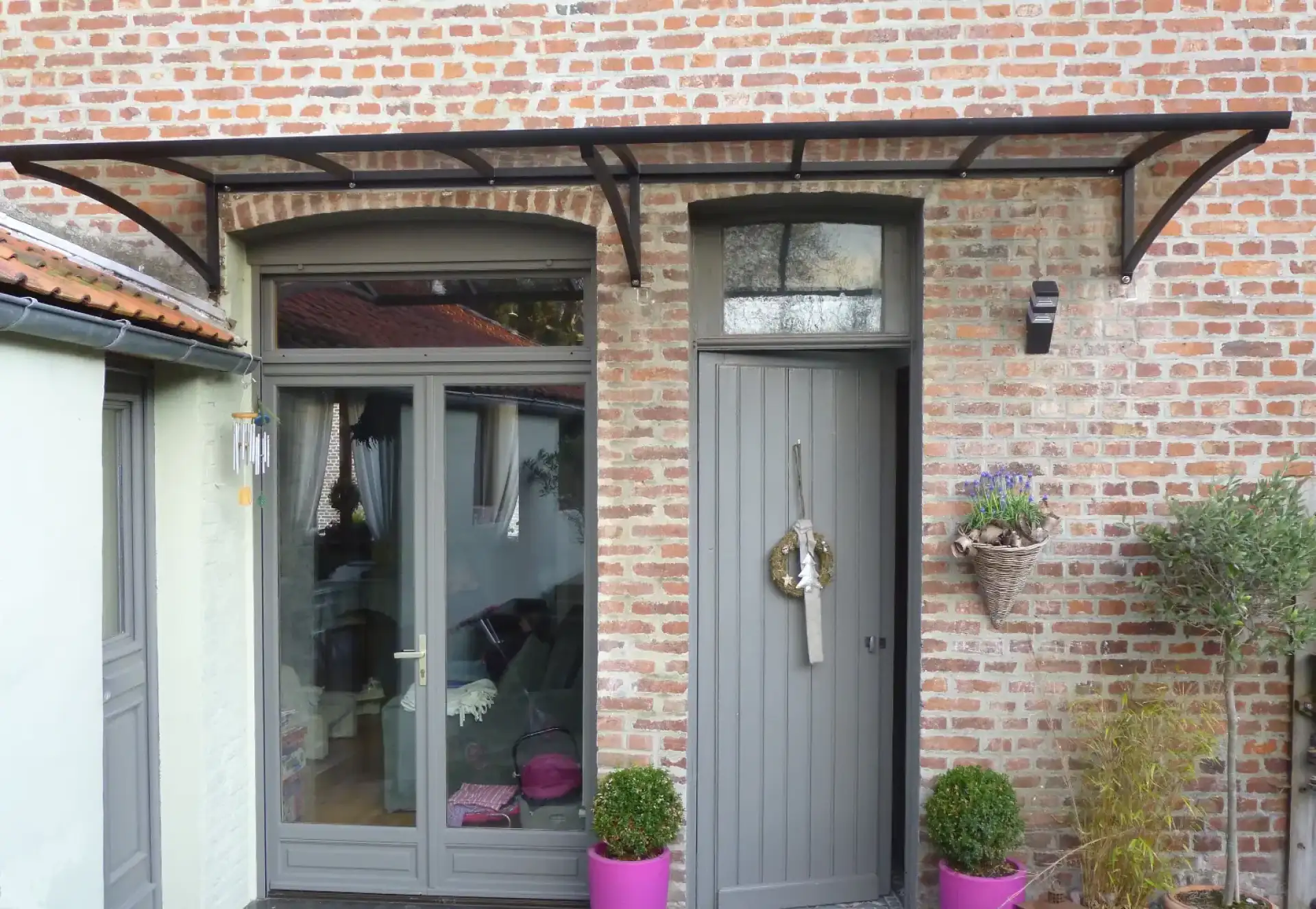 Entrée de maison en briques avec auvent moderne noir, double porte vitrée et porte grise, décorée de plantes et d'une couronne.