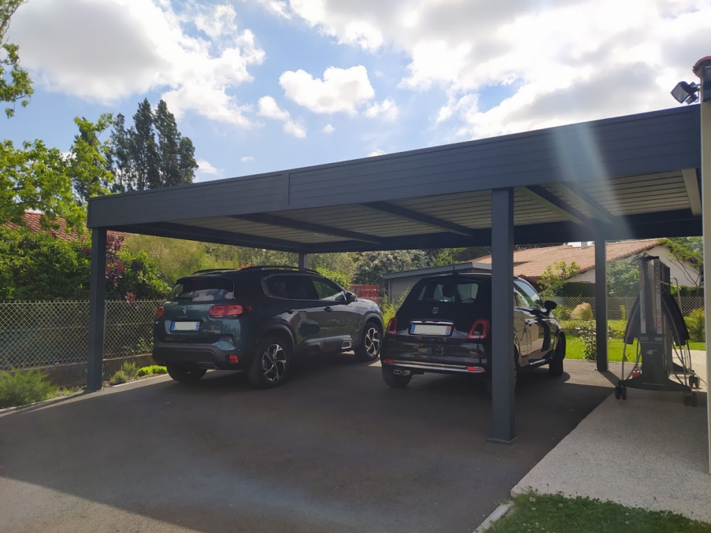 Carport moderne gris couvrant un SUV vert et une citadine noire. Jardin et clôture, ciel bleu nuageux.