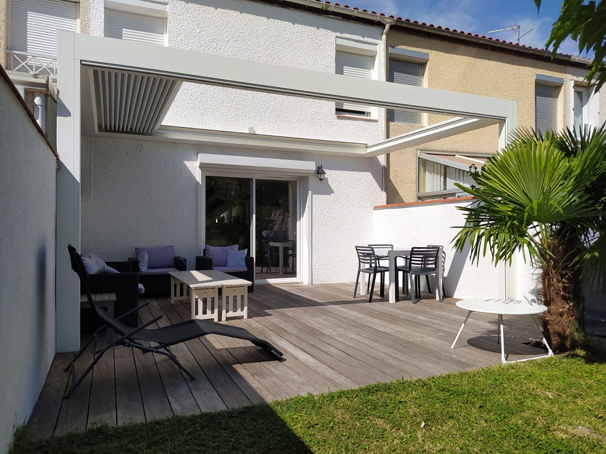 Terrasse en bois avec pergola blanche à lames orientables, salon de jardin sombre et table à manger.