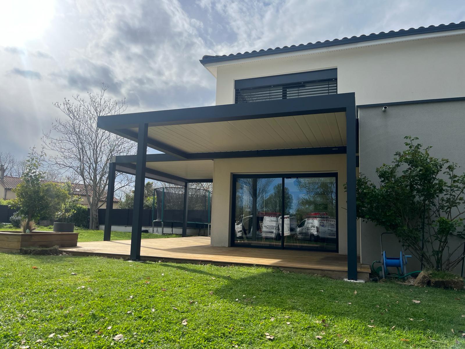 Maison moderne beige et grise avec pergola noire, grande terrasse bois et jardin arboré avec trampoline.