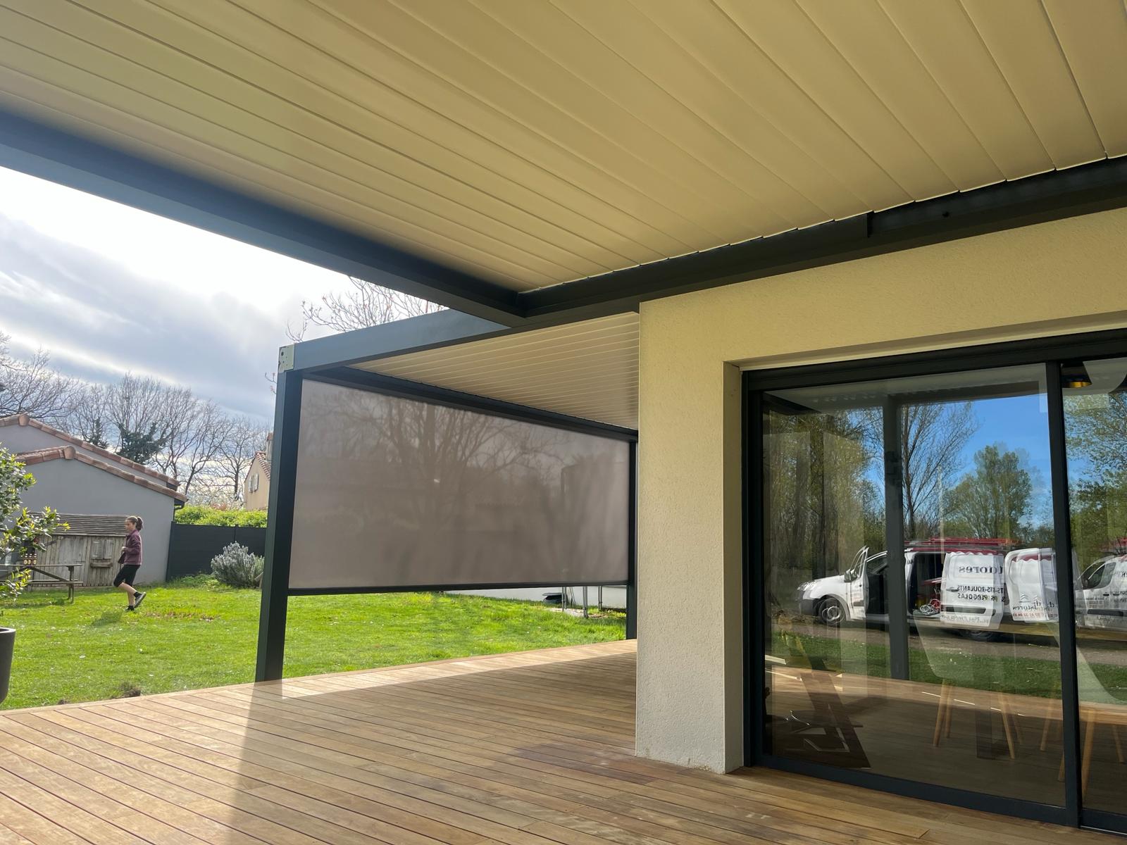 Pergola bioclimatique sur terrasse bois, avec store latéral gris baissé. Personne marche dans jardin. Reflets de vans blancs sur baie vitrée.