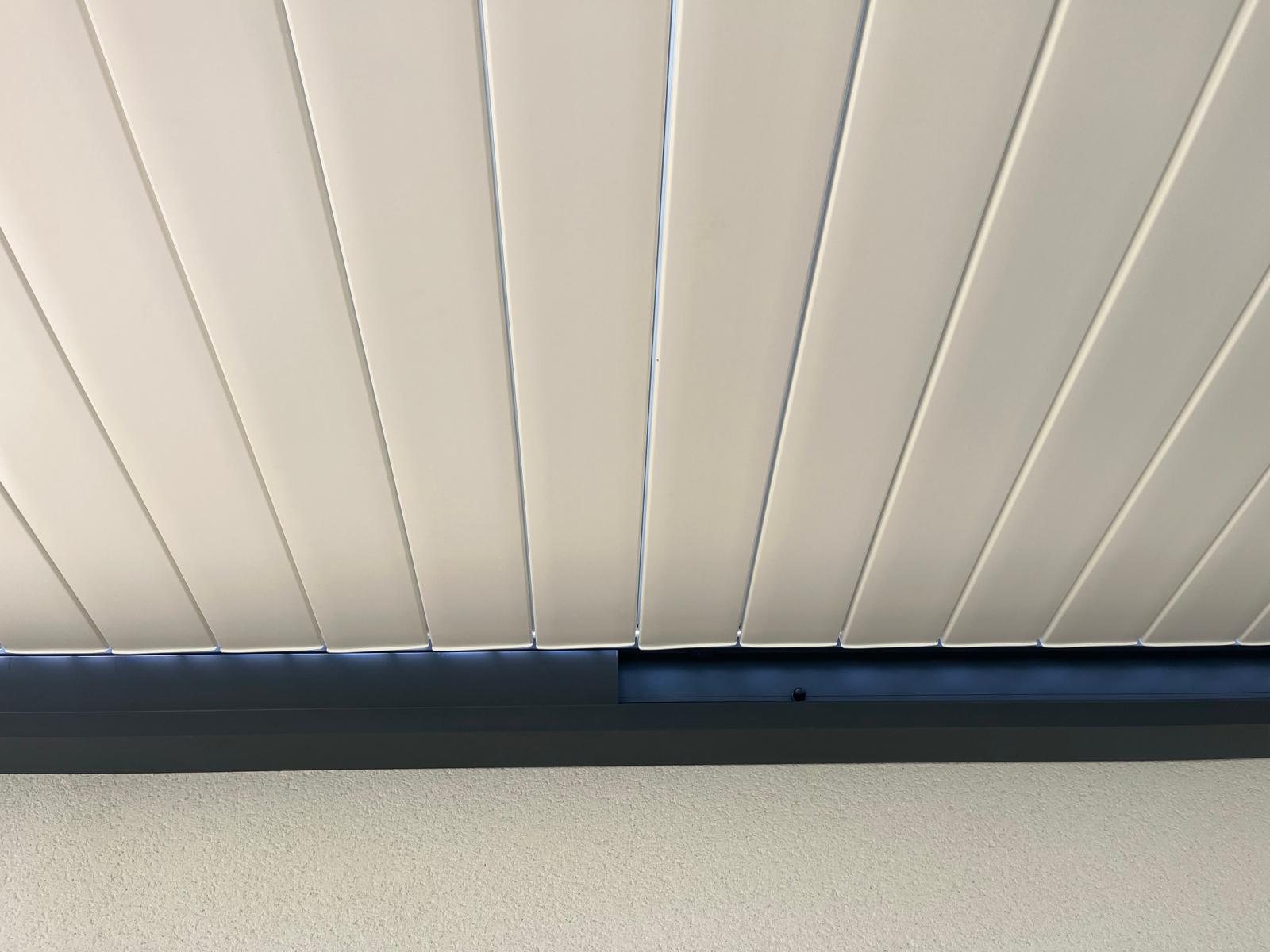 Toit de pergola blanc à lames parallèles légèrement ouvertes. Poutre gris foncé avec éclairage LED. Mur blanc texturé.