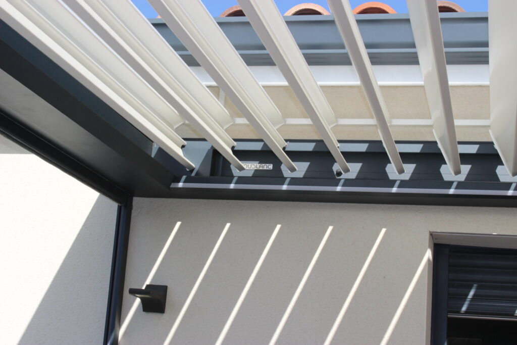 Pergola blanche à lames orientables, ombre portée sur mur texturé, éclairage et store, inscription OUDBLANC visible.