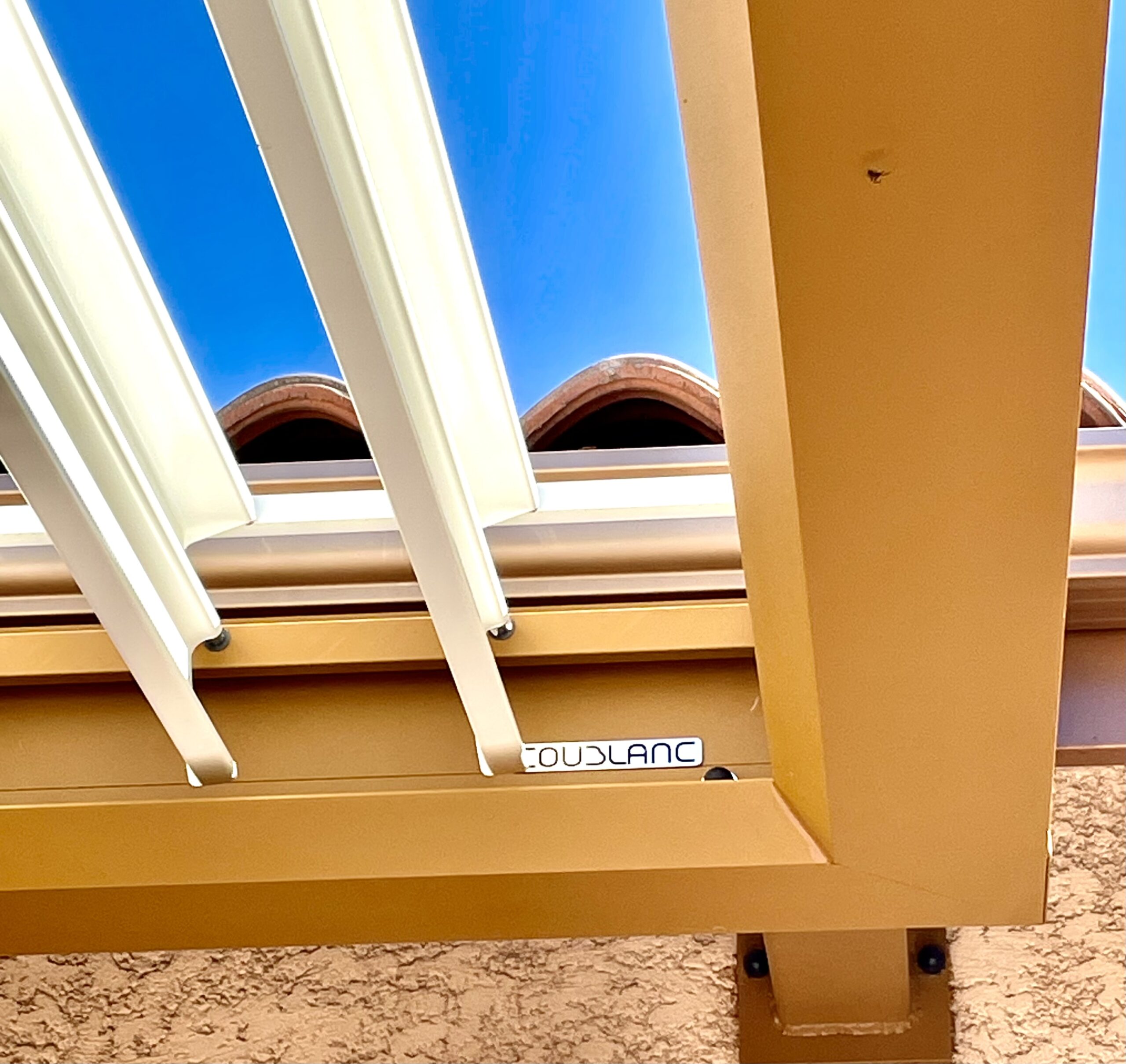 Structure de pergola beige avec louvers blancs, vue en contre-plongée sur le ciel bleu. Logo COUDBLANC visible sur la poutre.