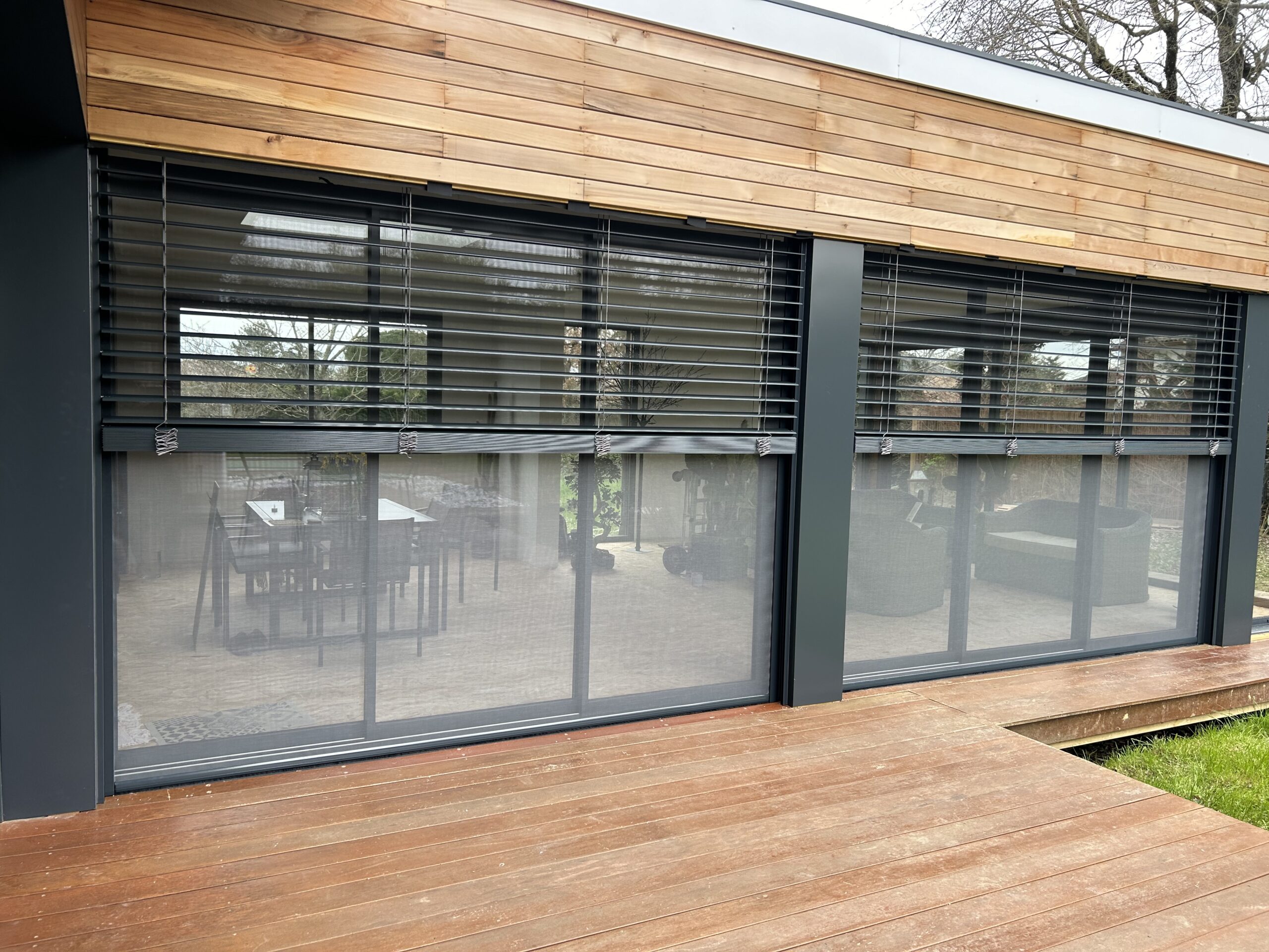 Façade moderne avec bardage bois, grandes fenêtres équipées de stores extérieurs foncés et de moustiquaires sur une terrasse en bois.