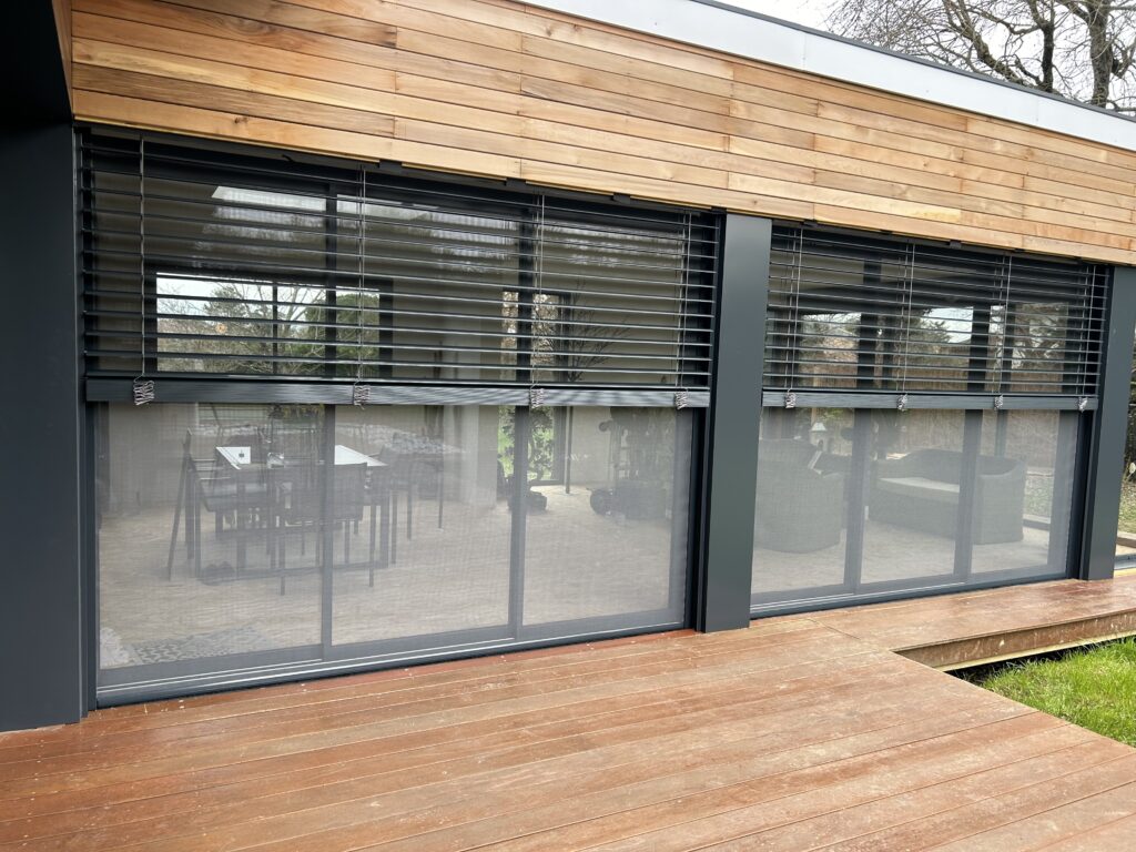 Façade bois moderne, stores extérieurs gris (persiennes, écrans), grande baie vitrée donnant sur terrasse bois.