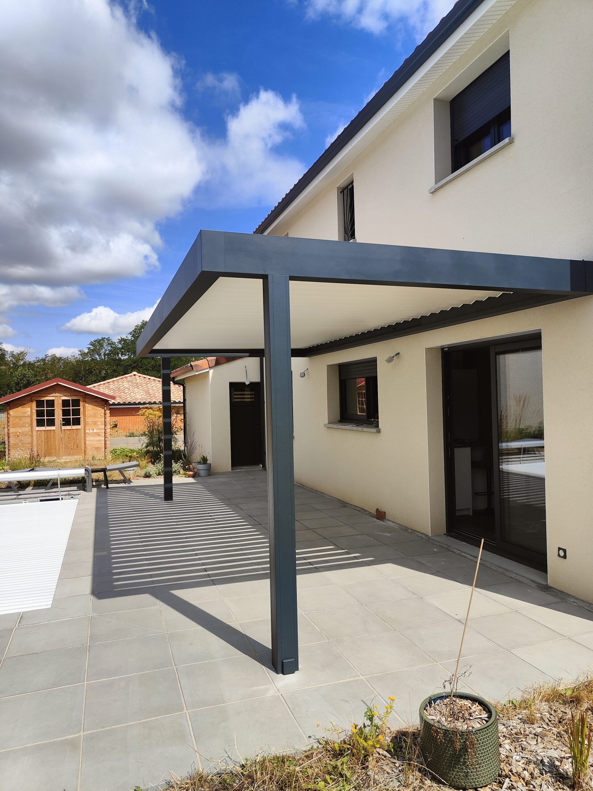 Maison moderne beige avec pergola grise sur terrasse carrelée, ombres portées. À gauche, piscine couverte et abri de jardin bois.