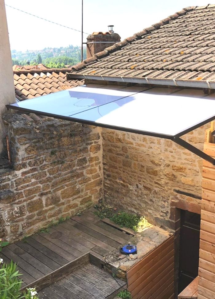 Canopée moderne transparente au-dessus d'une terrasse en bois adossée à un vieux mur en pierres. Vue sur un village.