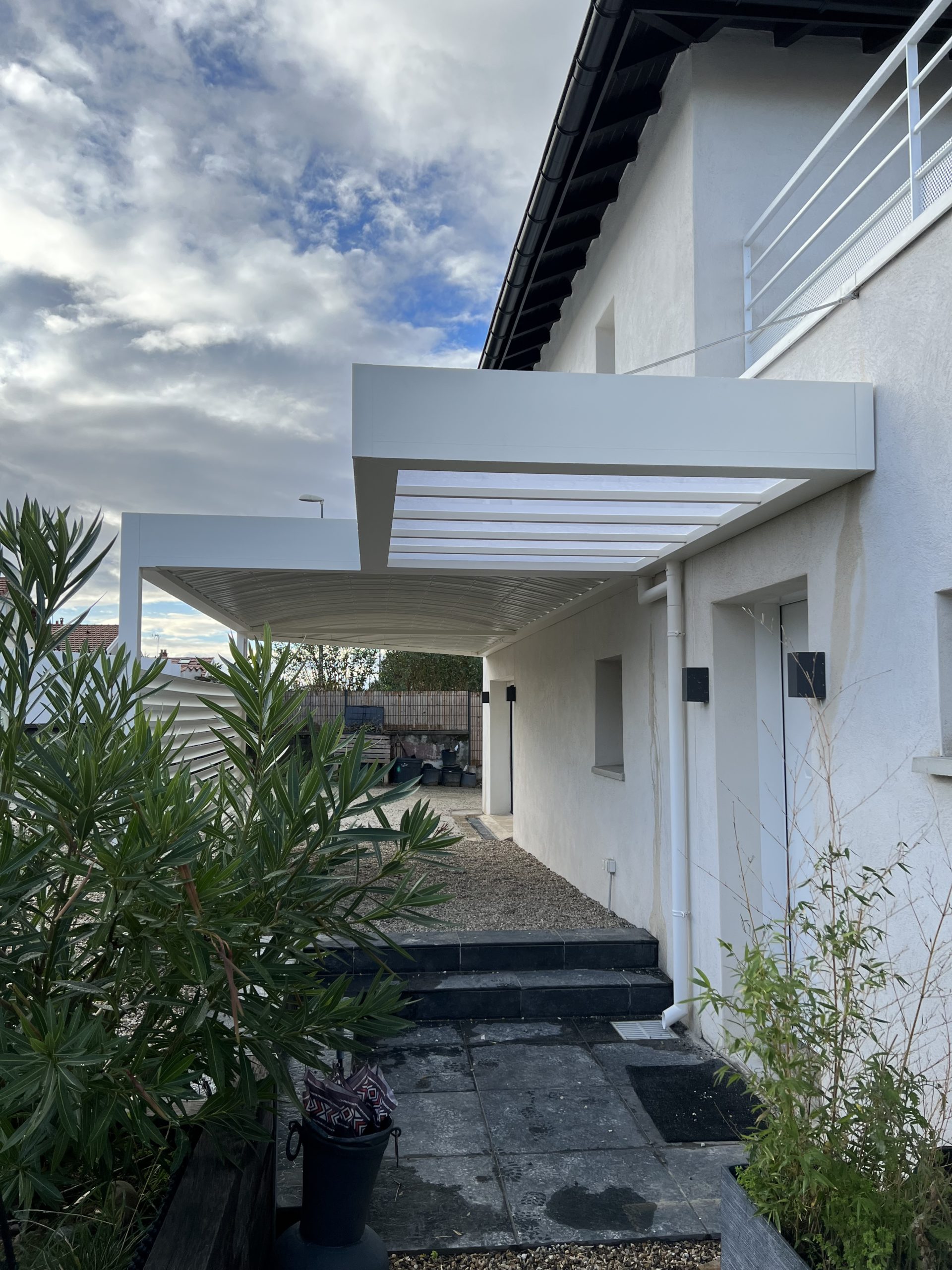 Maison moderne blanche avec pergola fixée, patio carrelé sombre, escalier et végétation luxuriante. Ciel nuageux.