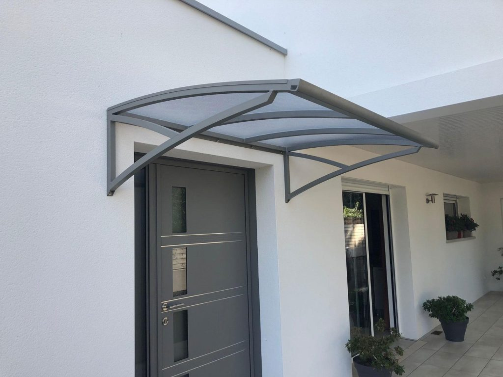 Entrée de maison moderne avec porte grise et auvent transparent incurvé soutenu par des bras gris.