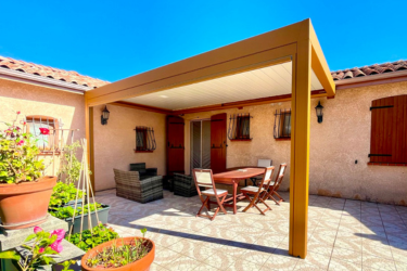 Pergola moderne beige sur terrasse ensoleillée avec table à manger et salon de jardin, devant une maison méditerranéenne.