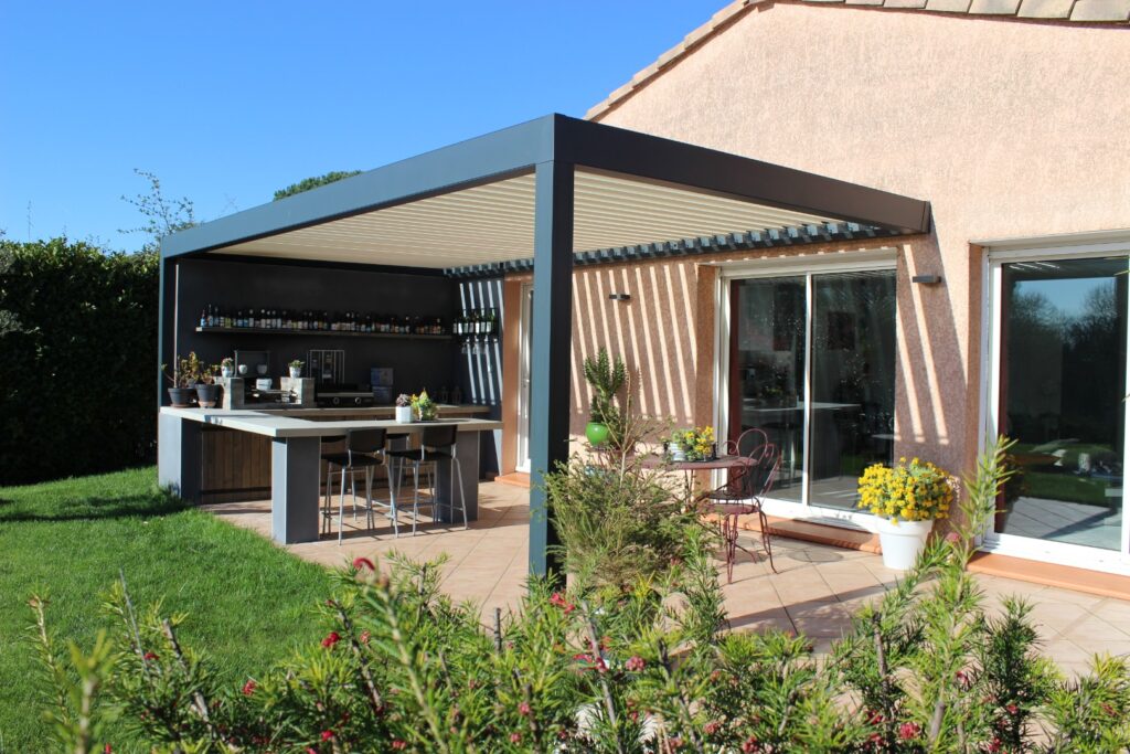 Pergola bioclimatique gris foncé sur mesure couvrant une cuisine extérieure moderne avec bar, tabourets et patio carrelé.