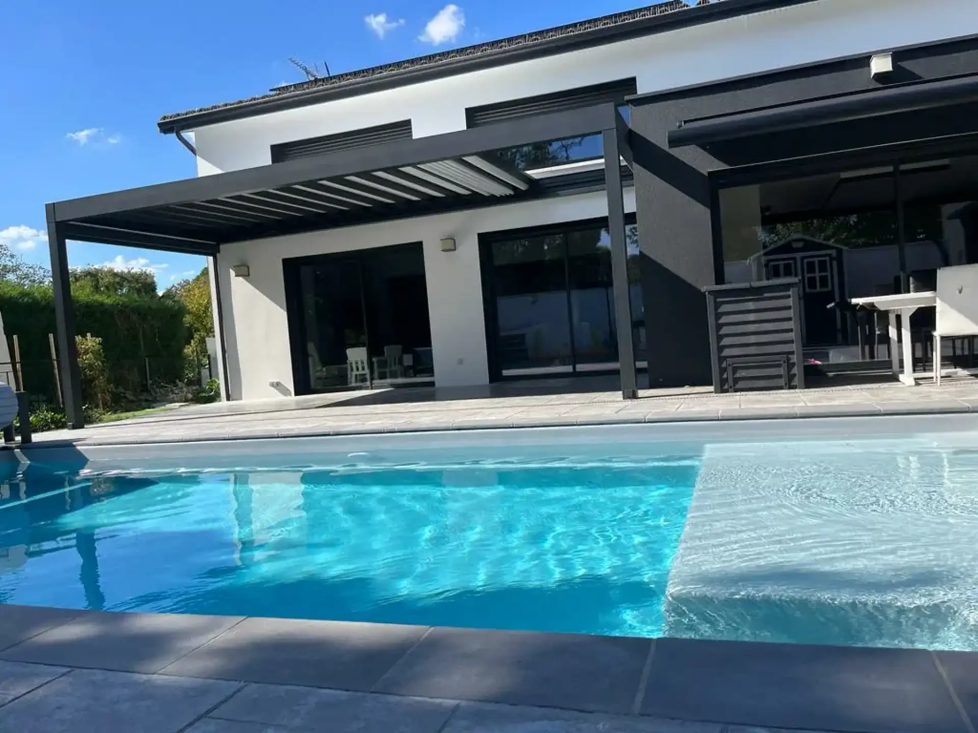 Maison moderne blanche/noire, piscine bleue cristalline, pergola bioclimatique sombre sur terrasse. Journée ensoleillée.