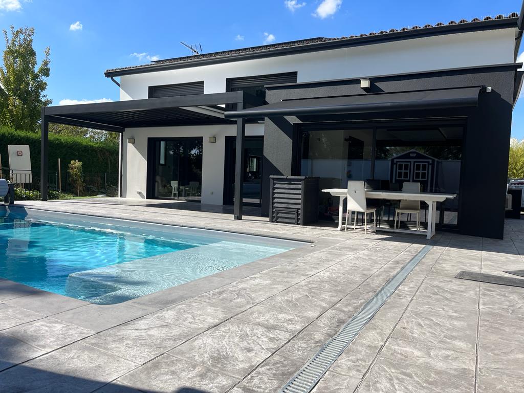 Maison moderne avec piscine bleue, grande terrasse grise pavée et pergola noire sous ciel bleu dégagé.