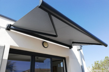 Store banne motorisé gris clair avec bras articulés noirs installé sur façade blanche au-dessus d'une baie vitrée.