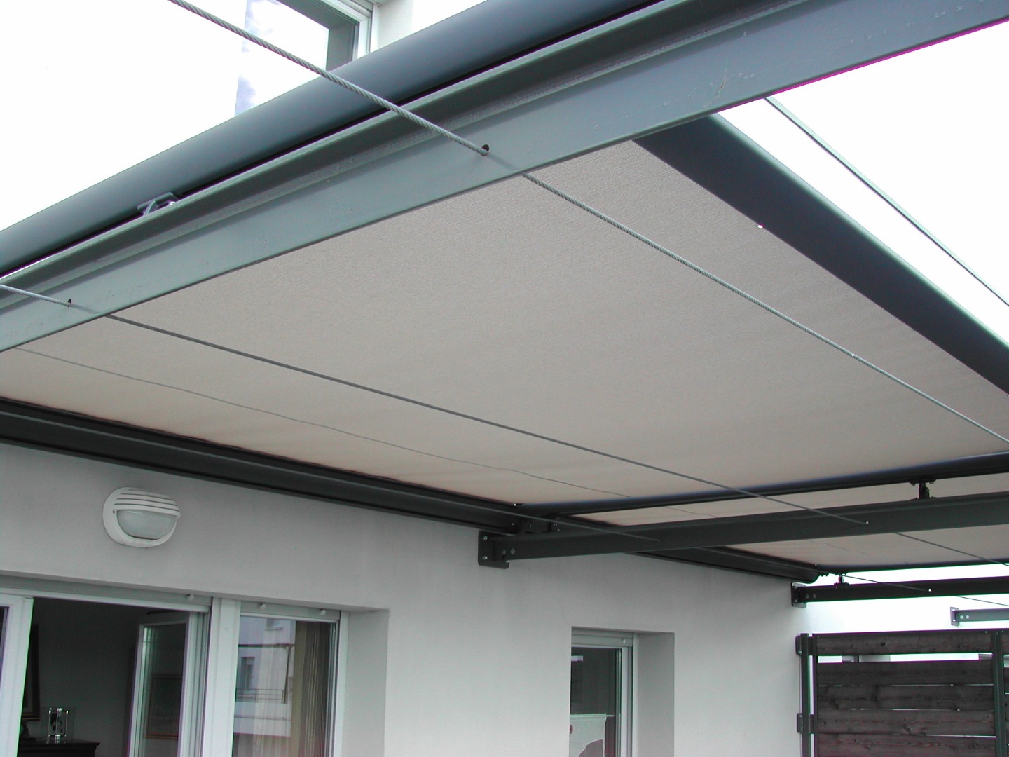 Store banne rétractable beige sur structure métallique foncée et câbles de guidage, installé au-dessus de fenêtres sur un mur blanc.