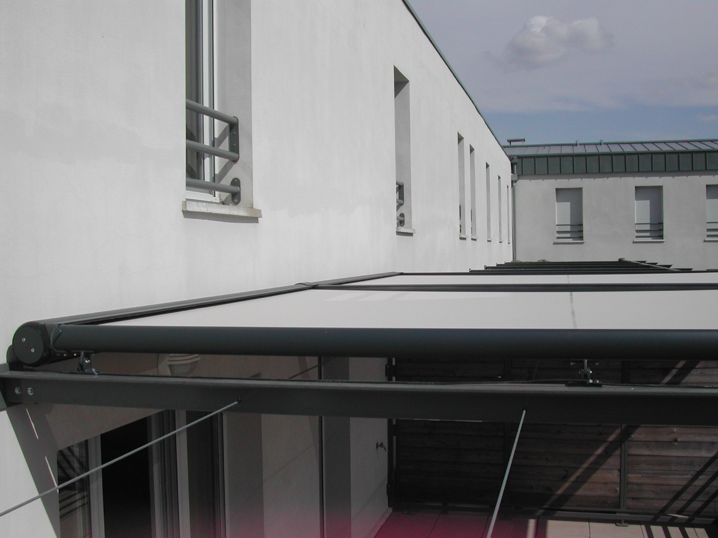 Pergola rétractable sur terrasse devant une façade blanche avec fenêtres grillagées. Bâtiment en arrière-plan.