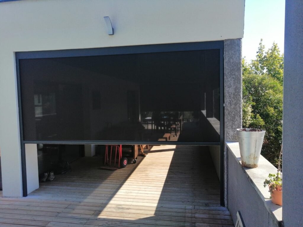 Store extérieur sombre semi-fermé sur terrasse en bois. Lumière du soleil forte projetant des ombres.