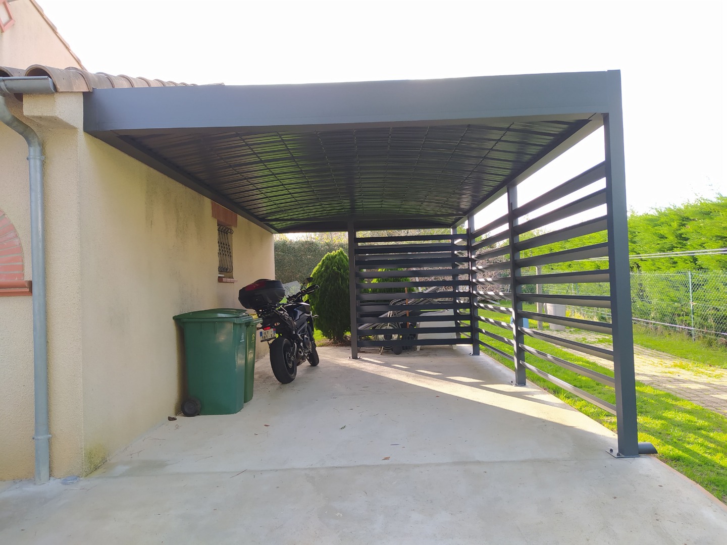 Carport noir adossé à une maison beige, abritant une moto noire et deux poubelles vertes. Mur latéral à lattes.