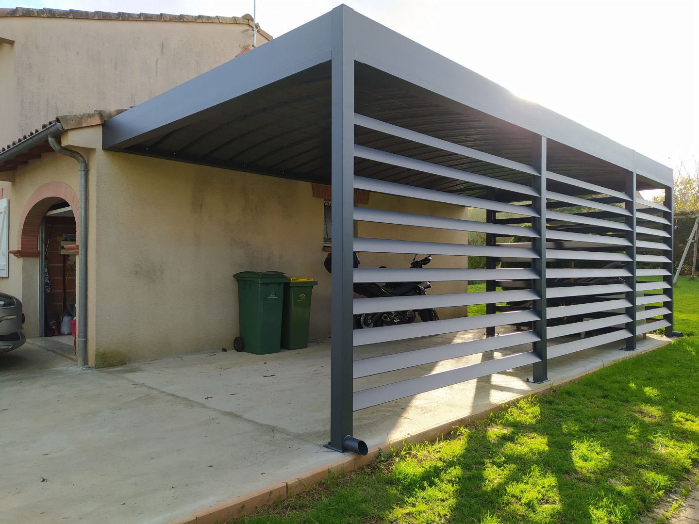 Carport moderne en métal gris adossé à une maison, avec parois à lattes. Couvre une allée bétonnée avec poubelles et motos garées.
