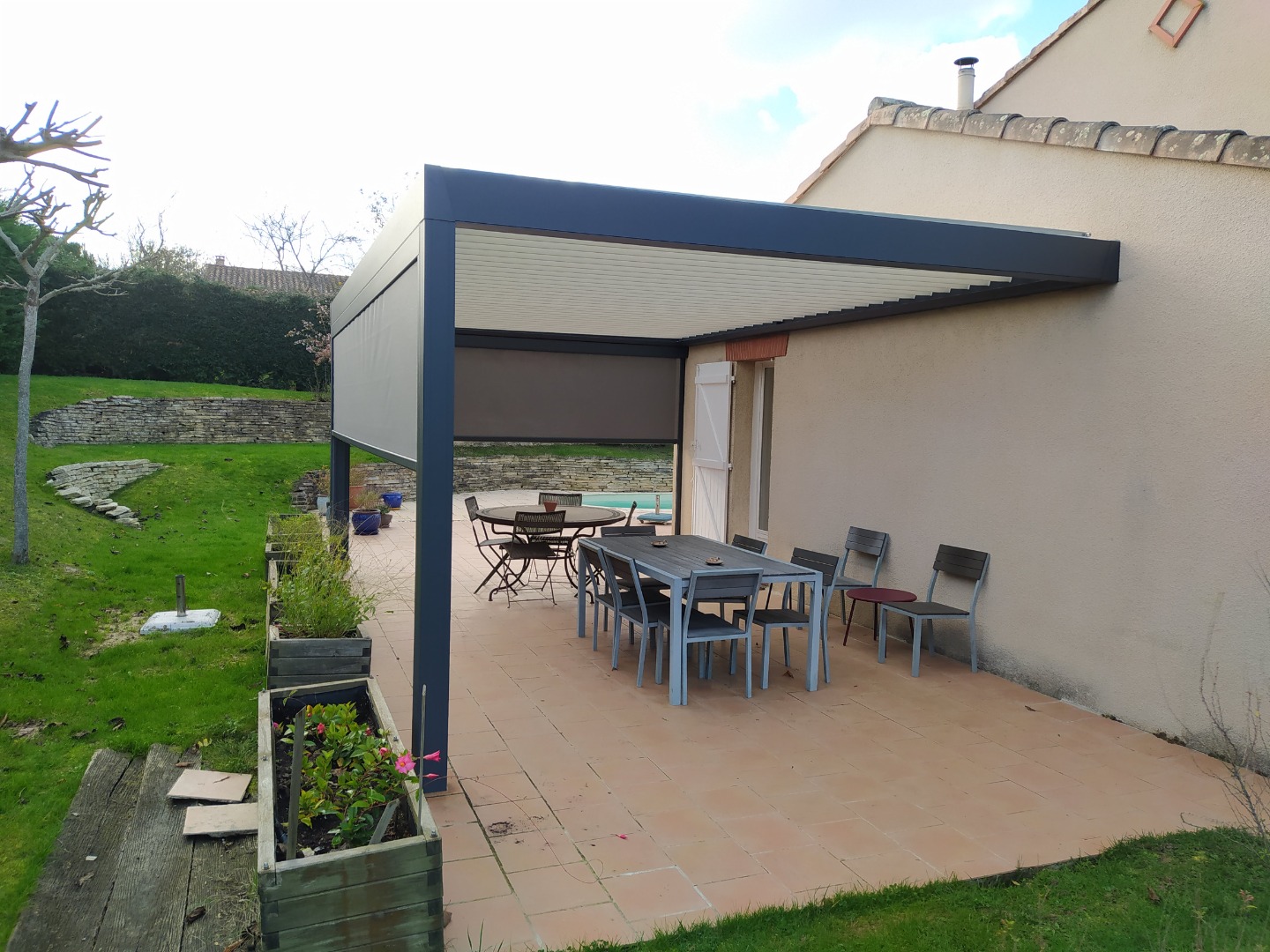 Pergola moderne anthracite sur terrasse en dalles terre cuite. Salon de jardin, store, jardin arboré et mur en pierre.