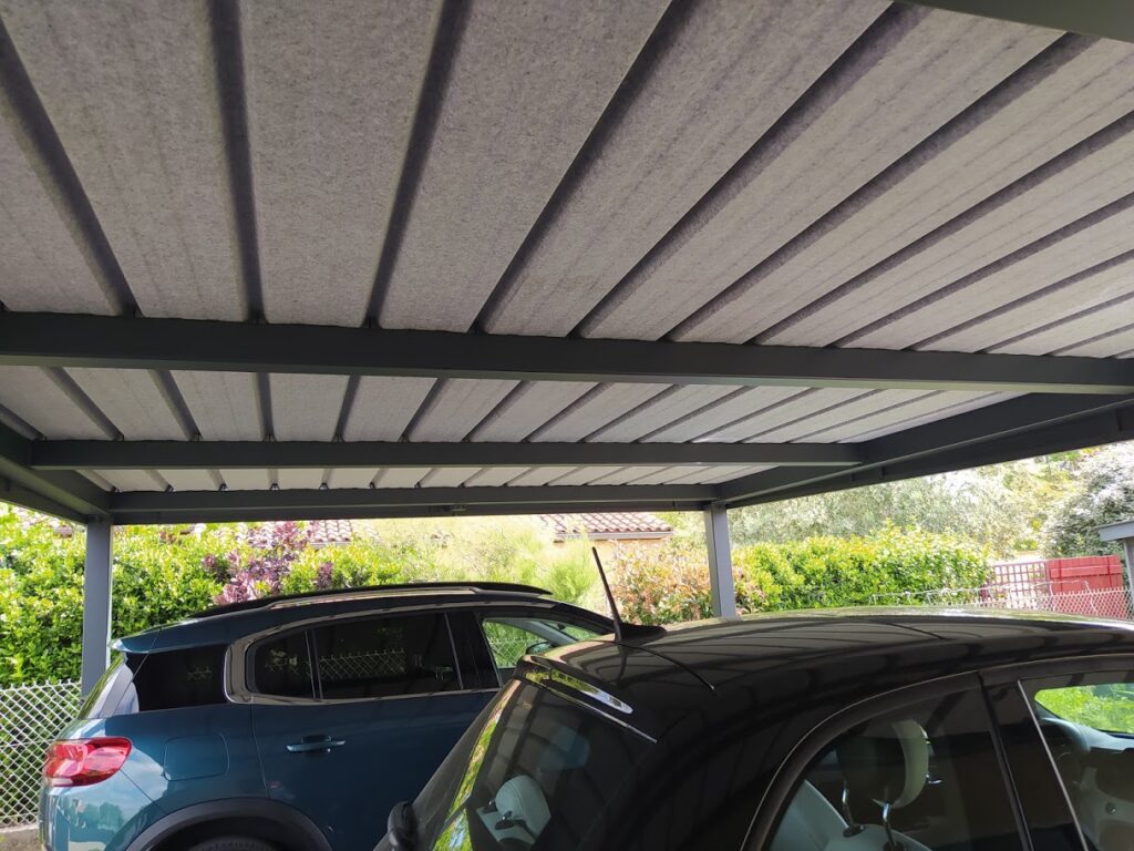 Deux voitures garées sous un carport en métal gris avec un toit ondulé gris texturé. De la végétation est visible en arrière-plan.
