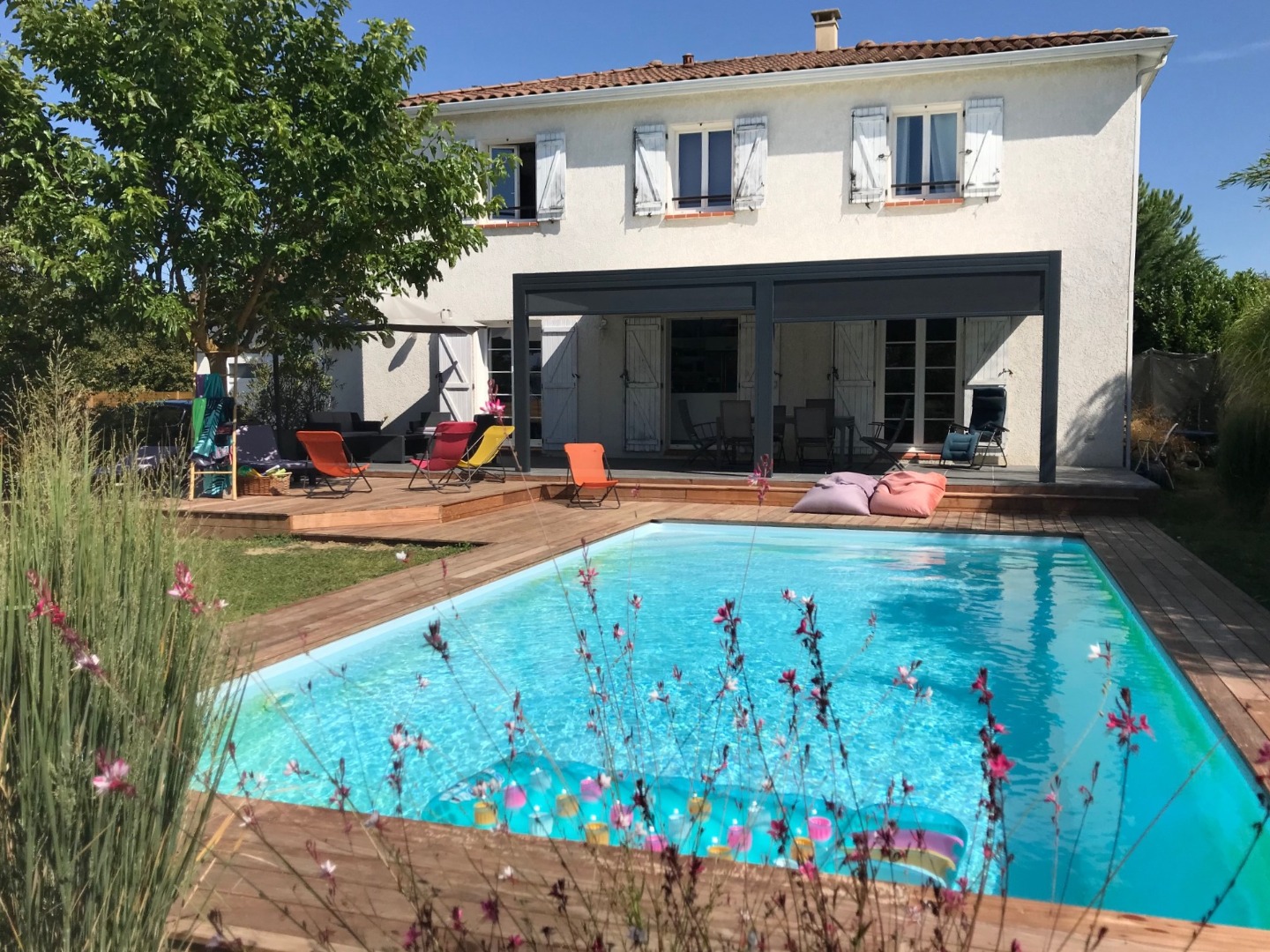 Maison blanche avec piscine, terrasse en bois, jardin ensoleillé et mobilier coloré au bord de l'eau
