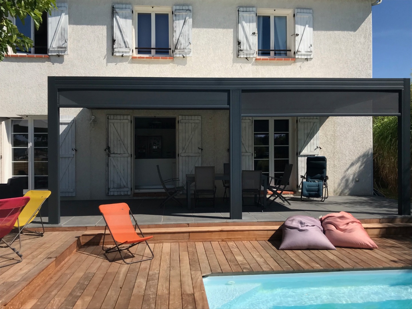 Maison blanche avec pergola grise couvrant une terrasse aménagée. Piscine avec deck bois, transats colorés et poufs.