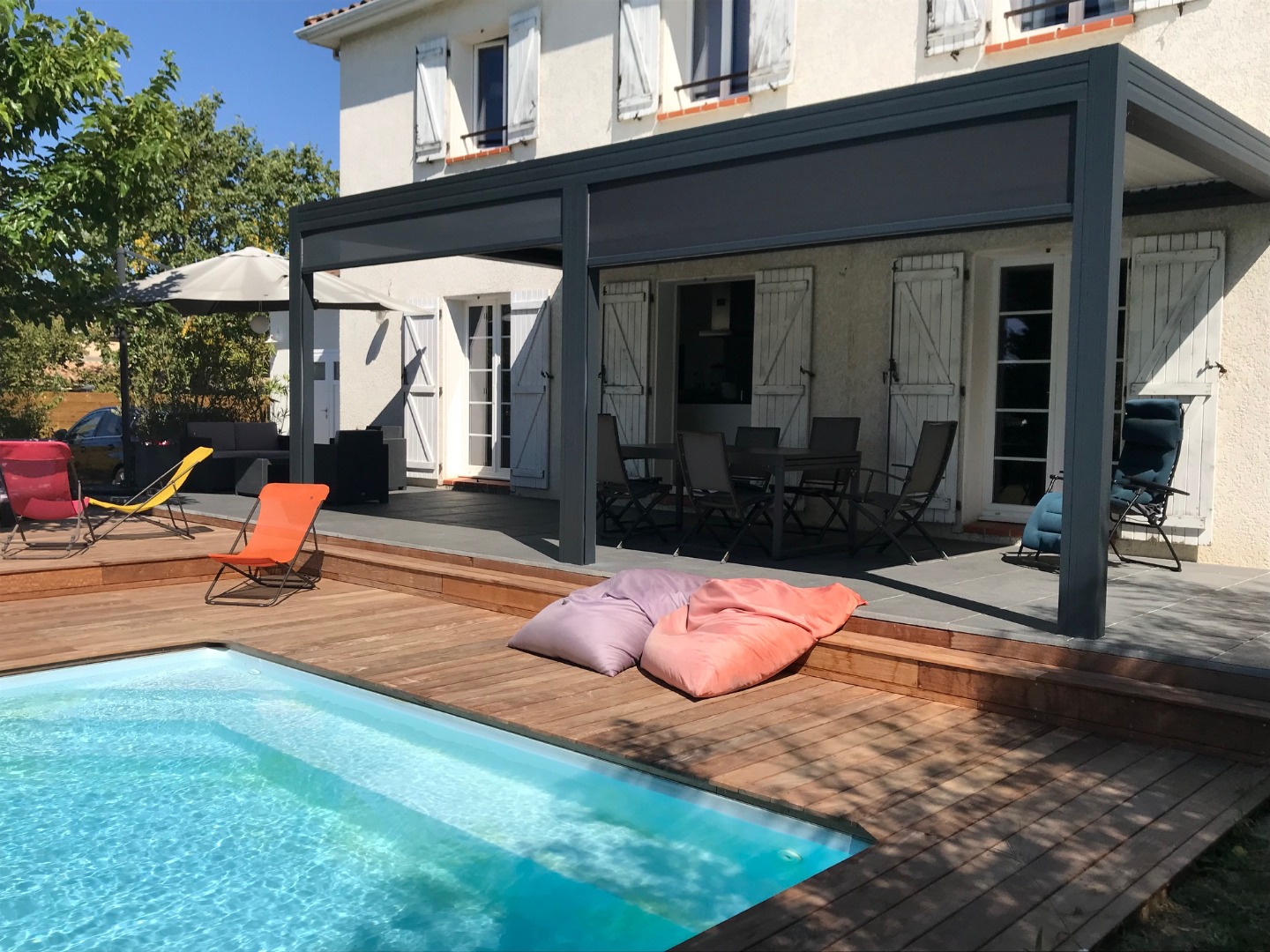 Piscine bleue, deck bois et terrasse carrelée sous une pergola moderne devant une maison. Mobilier d'extérieur coloré invite à la détente.