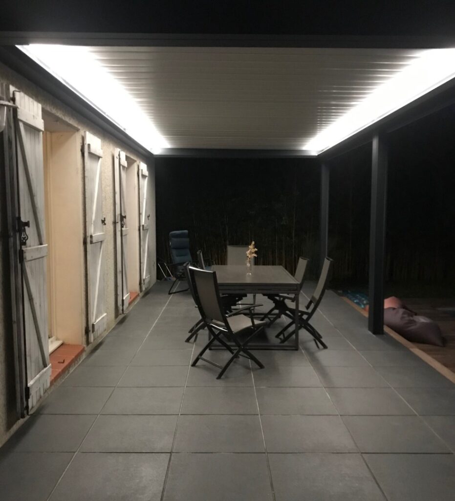 Terrasse extérieure carrelée la nuit. Pergola éclairée par LED au-dessus d'une table de jardin et chaises. Volets sur la gauche, fond sombre.