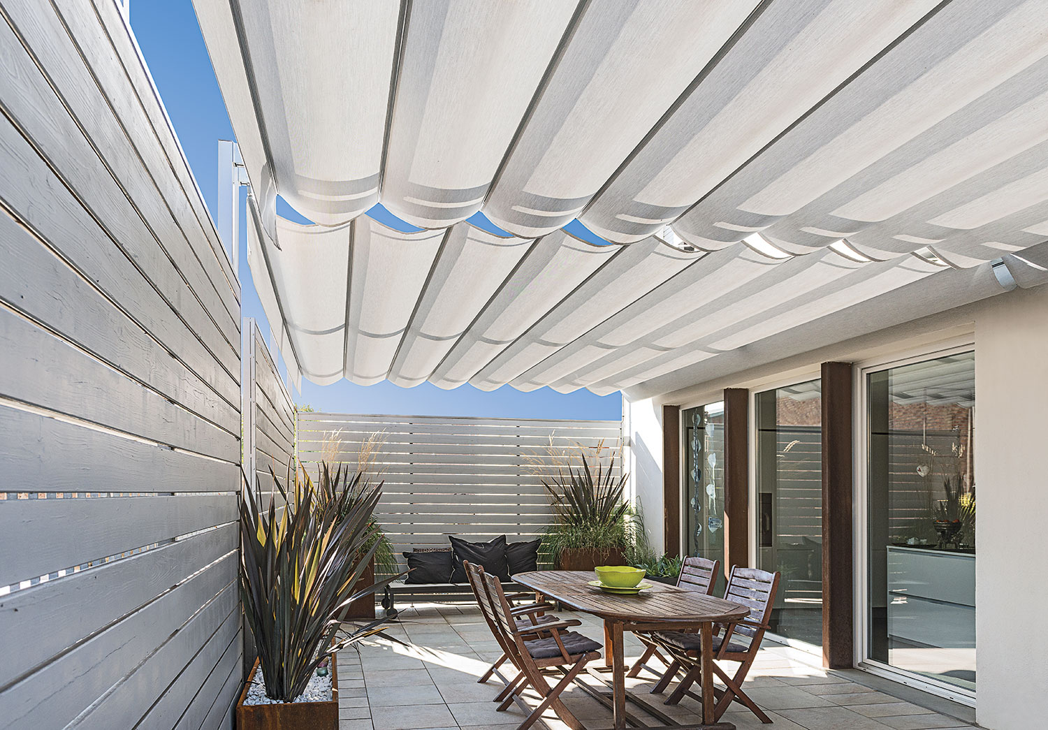 Terrasse moderne avec pergola en toile rétractable blanche, mobilier de jardin en bois et clôture ajourée. Espace extérieur design.