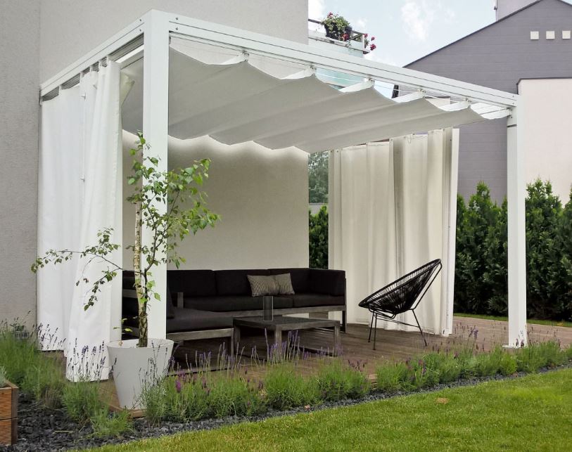 Pergola blanche moderne avec toit rétractable et rideaux sur terrasse bois. Mobilier sombre, plantes et gravier entourent l'espace.
