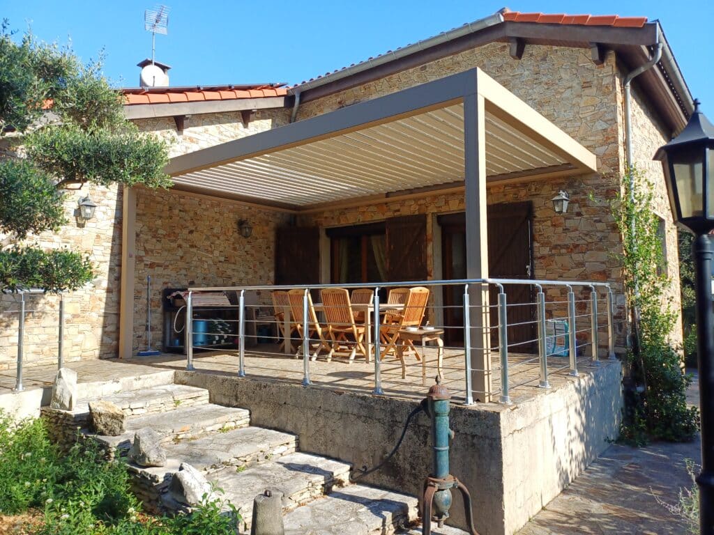 Grande terrasse de maison en pierre sous une pergola bioclimatique marron clair, avec balustrade inox et escalier extérieur.