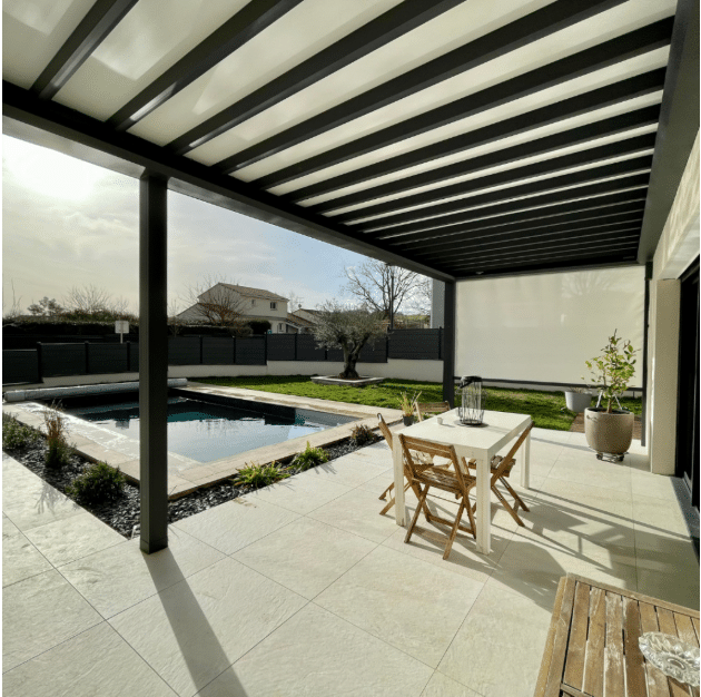 Terrasse moderne avec pergola anthracite couvrant un espace repas et la piscine. Carrelage clair, jardin arboré et store latéral rétractable.